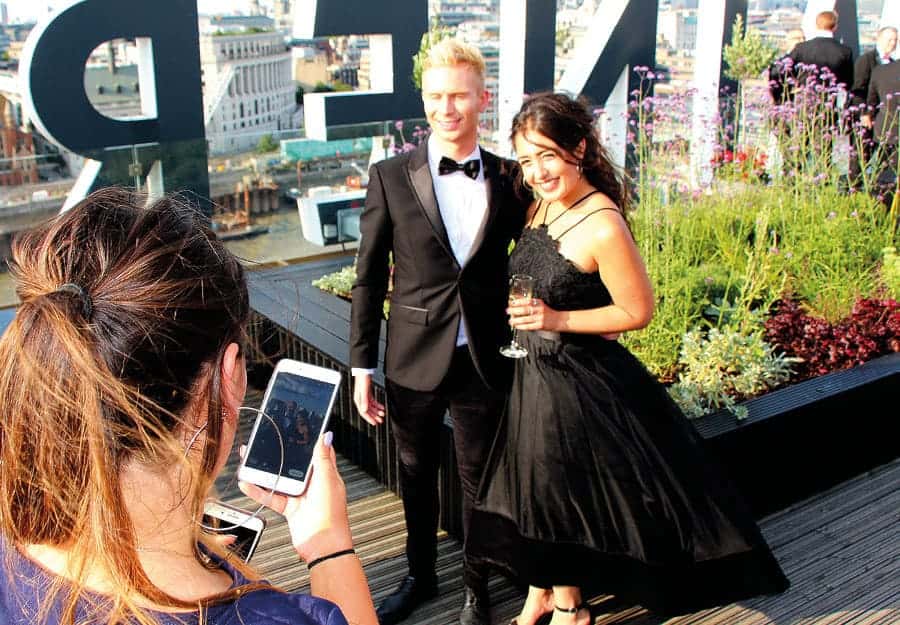 Twee mensen, gekleed in galakleding, staan op een dakterras. Een derde persoon neemt een foto van hen op een telefoon. Het weer is zonnig.