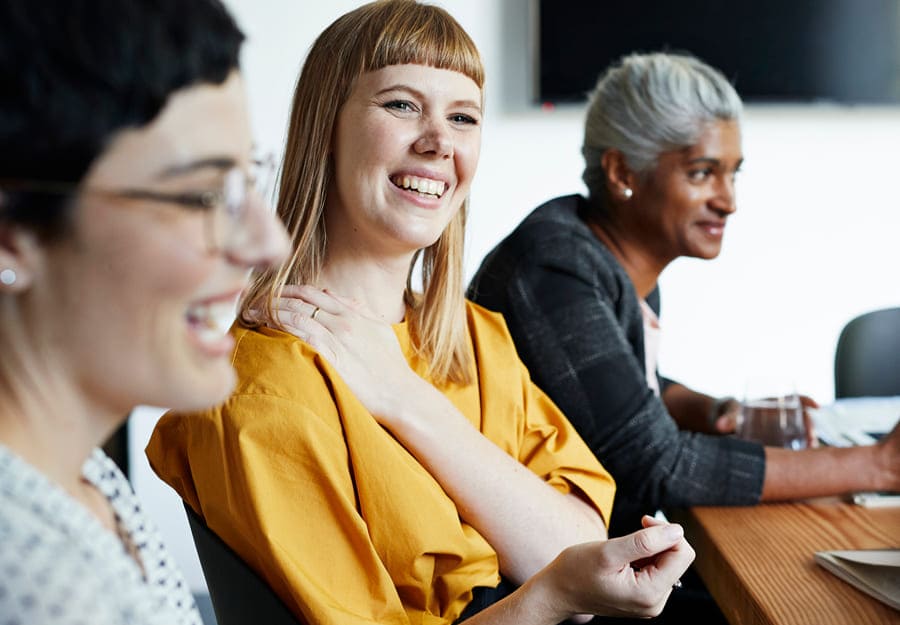 Drie vrouwen zitten aan een tafel en glimlachen. De focus ligt op de vrouw in het midden - ze draagt een felgeel shirt en glimlacht terwijl ze haar linkerhand comfortabel op haar rechterschouder laat rusten. 