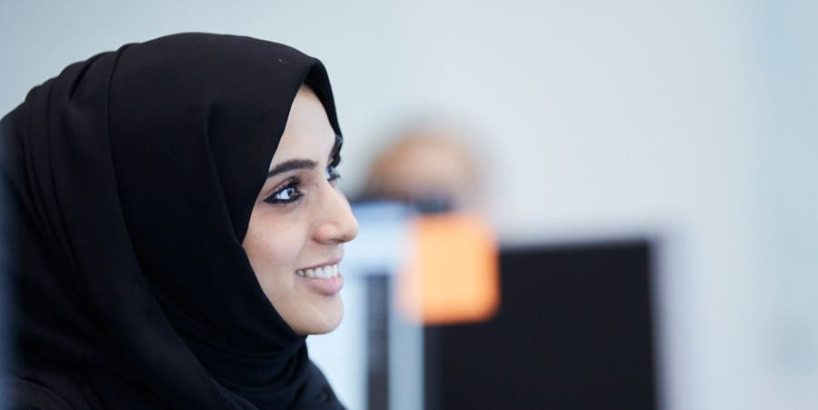 Een vrouw met een zwarte hoofddoek glimlacht en richt haar blik weg van de camera. De foto is een zijwaartse hoek van haar gezicht. 