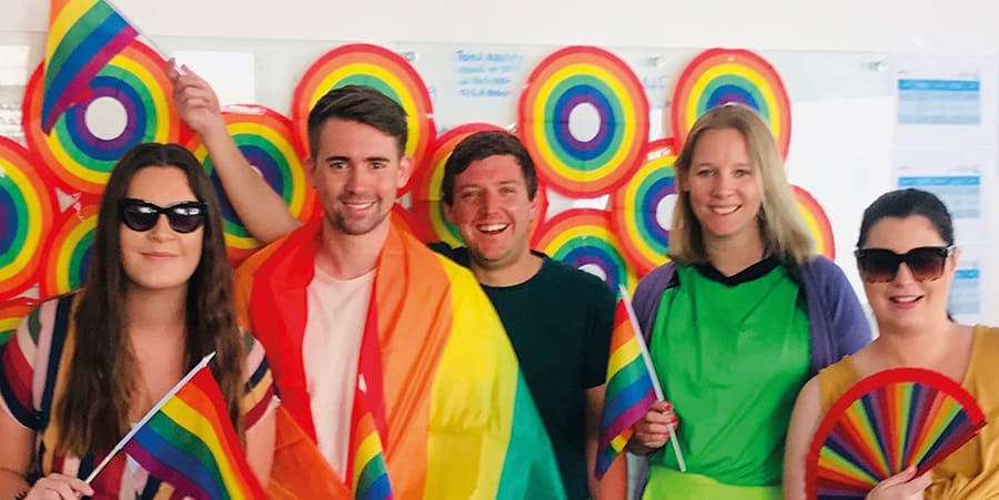 Vijf mensen lachen naar de camera, en zwaaien met regenboogkleurige vlaggen en waaiers