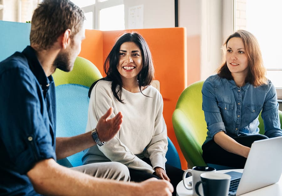 Drie personen zitten aan een tafel met een laptop en enkele mokken. Het trio is in gesprek.